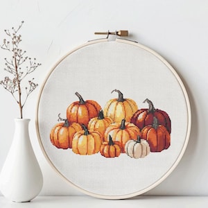 May include: A cross-stitch embroidery of pumpkins in various shades of orange, yellow, and brown. The pumpkins are arranged in a cluster, with a white pumpkin in the front. The embroidery is set in a wooden hoop.