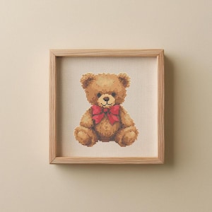May include: A framed cross-stitch of a teddy bear. The bear is brown with a red bow tie. The artwork is displayed in a light-colored wooden frame, set against a neutral background.
