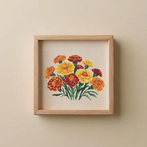 Peut inclure: Une broderie au point de croix encadrée représentant des fleurs de souci aux tons jaune, orange et rouge, avec des feuilles vertes. L'œuvre est présentée dans un cadre en bois naturel. C'est un objet décoratif carré.