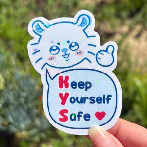 May include: A sticker featuring a cartoon cat with a thumbs-up gesture. The cat is light blue with glitter accents. A speech bubble contains the text "Keep Yourself Safe" in red and blue, with a heart symbol.