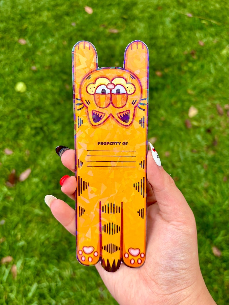 May include: Orange Garfield-themed phone case with a cartoon design. The case features Garfield's face at the top and his body extending down. The text "PROPERTY OF" is printed on the case, with lines for personalization. The case is held in a hand.