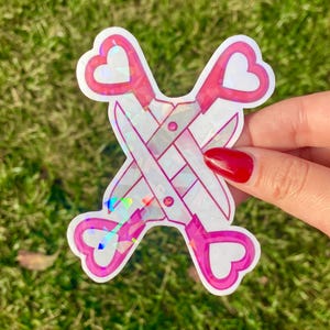 May include: A sticker of a pair of scissors with pink heart-shaped handles. The scissors are crossed and the blades are iridescent.