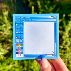 May include: A blue sticky note with a computer-style paint program interface design. The note features the text "untitled - Paint" at the top, with a blank white space in the center, and a color palette on the left side. The note is held by a hand.