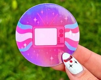 May include: A round sticker with a pink, blue, and white transgender pride flag design. A pink retro television with a white screen is in the center of the flag. The sticker has a holographic finish and sparkles.