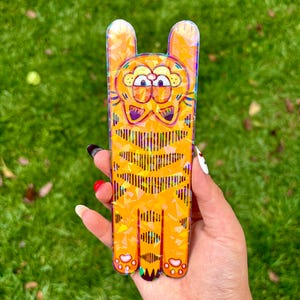 May include: A rectangular, orange Garfield-shaped phone case with a cartoon design. The case features a yellow and orange color scheme with a striped pattern on the body and ears. The case is held against a green grassy background.
