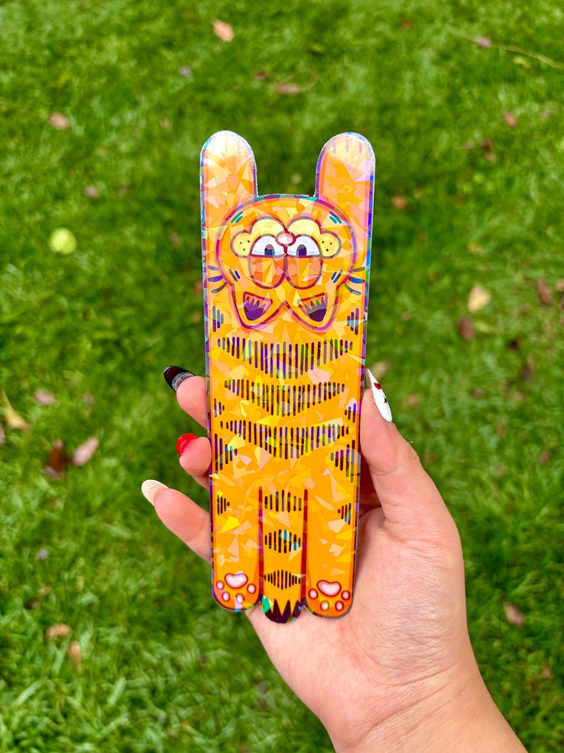 May include: A rectangular, orange Garfield-shaped phone case with a cartoon design. The case features a yellow and orange color scheme with a striped pattern on the body and ears. The case is held against a green grassy background.