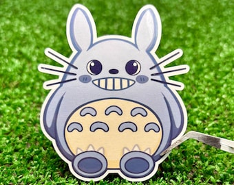 May include: A die-cut sticker featuring a cartoon creature with a round, light blue body, tall ears, and long whiskers. The character has a wide, toothy grin, dark eyes, and a cream-coloured belly with dark blue chevron markings and two large feet.