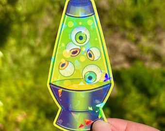 May include: A holographic sticker in the shape of a lava lamp, filled with green liquid and floating eyeballs. The sticker has a yellow outline and a dark blue base and top. The sticker is held by a hand against a blurred green background.