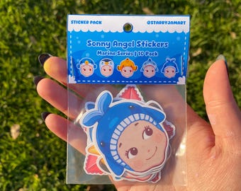 May include: A pack of Sonny Angel stickers from the Marine Series, featuring cute cartoon characters in marine-themed outfits. The stickers are in a clear plastic bag, held in a hand, with a green grassy background.