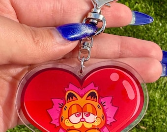 Può includere: Un portachiavi in ​​acrilico a forma di cuore rosso con un'immagine di cartone animato di Garfield all'interno. Garfield è arancione con occhi neri e un naso rosa. Indossa un fiore rosa sulla testa e ha un cuore rosa negli occhi.
