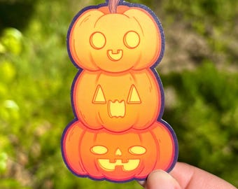 May include: A stacked pumpkin sticker with a purple border. The sticker features three orange pumpkins with carved faces, each with a unique expression. The top pumpkin has a smiling face, the middle one has a surprised face, and the bottom one has a jack-o'-lantern face.