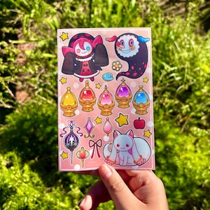 May include: A sticker sheet featuring various cartoon illustrations. The designs include a character in a red and black outfit, a floating ghost-like figure, and several jewel-like orbs. Other images include a white cat with pink accents, stars, and a bow.