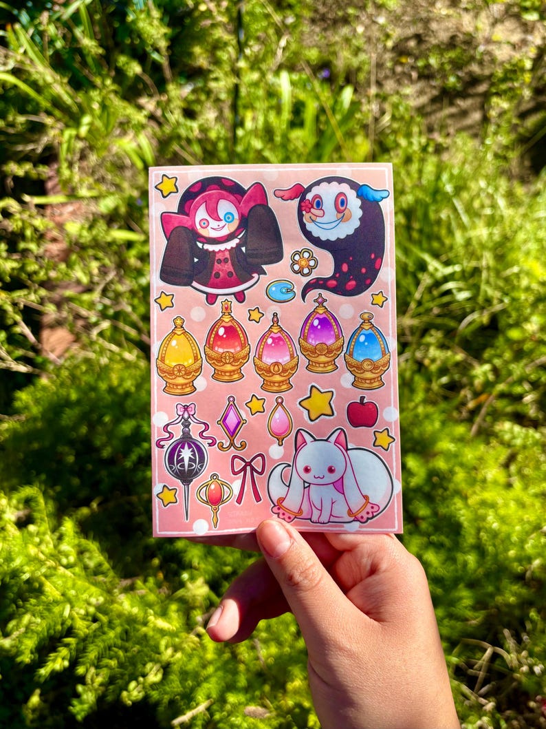 May include: A sticker sheet featuring various cartoon illustrations. The designs include a character in a red and black outfit, a floating ghost-like figure, and several jewel-like orbs. Other images include a white cat with pink accents, stars, and a bow.