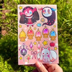 May include: A sticker sheet featuring colorful anime-style illustrations. The designs include various characters, magical gems, stars, and a white cat-like creature with pink accents. The background is a light pink color.