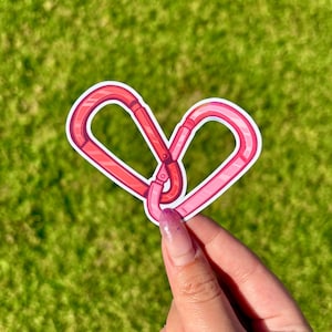 May include: A heart-shaped sticker formed from two interlocking carabiners. The carabiners are in shades of pink and red, with a white border. The sticker is held against a backdrop of green grass.