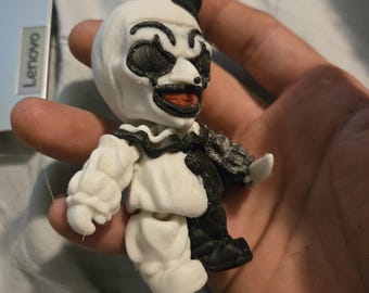 May include: A 3D-printed figurine of a clown with a black top hat, white face, and black and red facial details. The clown has a black and white outfit. The figurine is held in a hand.