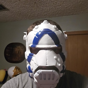May include: A white full-face helmet featuring bold blue geometric accents and a segmented design. The helmet includes a prominent chin guard with vertical ventilation slots, raised circular details, and side protrusions, covering the head and face.