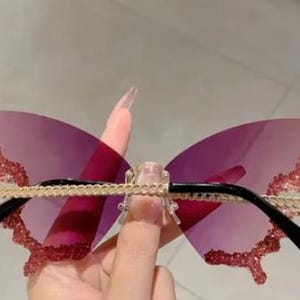 May include: Butterfly-shaped sunglasses with a pink gradient lens. The frames have a gold chain design across the top and black arms. The edges of the butterfly wings are embellished with pink glitter.