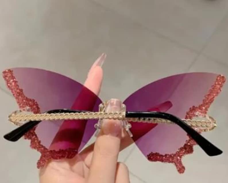May include: Butterfly-shaped sunglasses with a pink gradient lens. The frames have a gold chain design across the top and black arms. The edges of the butterfly wings are embellished with pink glitter.