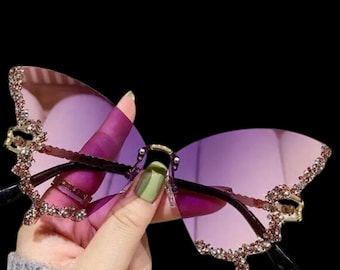 Puede incluir: Gafas de sol con forma de mariposa con lentes moradas y borde decorativo de pedrería. Las monturas son de color marrón oscuro con detalles dorados. Diseño único y elegante.
