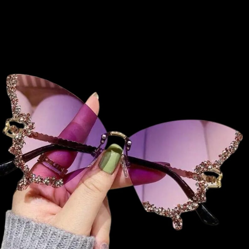 May include: Butterfly-shaped sunglasses with purple lenses and a decorative rhinestone border. The frames are dark brown with gold accents. The sunglasses have a unique, elegant design.