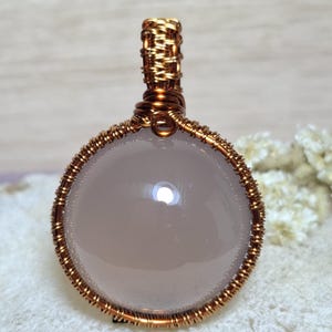 May include: A round, pale pink gemstone pendant wrapped in copper wire. The wire is intricately woven around the stone and forms a loop at the top for a chain. The pendant is a sphere, and the wire has a textured appearance.