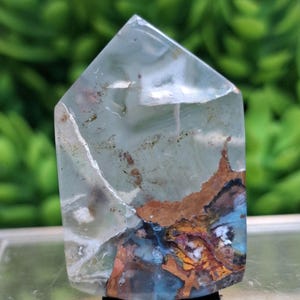 May include: A clear, polished crystal point with internal inclusions of brown, blue, and orange hues. The crystal has a flat base and a pointed top. The background is blurred green.