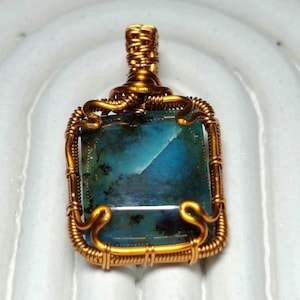 Può includere: Un ciondolo fatto a mano con una pietra preziosa rettangolare blu-verde. La pietra è incastonata in una cornice di filo dorato con dettagli intricati. Il ciondolo ha un anello nella parte superiore per una catena.