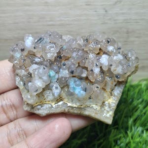 May include: A cluster of botryoidal chalcedony, showcasing a mix of translucent and opaque formations. The stone features a palette of pale gray, white, and hints of blue-green, with small black inclusions. The base is a light beige color.