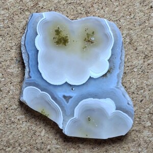 May include: A polished, irregularly shaped agate stone with a cloud-like pattern. The stone features concentric bands of white, light blue, and gray, with areas of green and brown inclusions. The overall shape is reminiscent of a stylized flower or cloud.