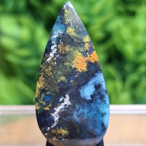 May include: A teardrop-shaped gemstone with a polished surface. The stone features a blend of colors, including deep blues, blacks, yellows, and whites, creating a unique pattern. The gemstone is set against a blurred green background.