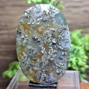 以下が含まれることがあります： 楕円形のモスアゲートの宝石。石は、緑、茶色、白の色が混ざり合い、苔のような模様が特徴です。宝石は透明なアクリルスタンドに展示されており、その自然な美しさを際立たせています。