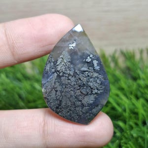 May include: A teardrop-shaped gemstone with a unique pattern. The stone features a mix of gray, black, and white hues, with a textured, almost landscape-like design. The gem is held between fingers against a blurred green background.