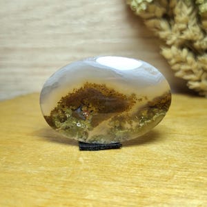 Pode incluir: Uma pedra de ágata polida, em forma oval, com um padrão de paisagem natural. A pedra apresenta faixas brancas e cinzentas translúcidas, com inclusões castanhas e verdes que se assemelham a uma paisagem em miniatura. É exibida num pequeno suporte preto.