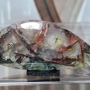 以下が含まれることがあります： 半透明で曇った白いベースに、濃い茶色、赤茶色、緑色の鉱物の内包物が全体に広がる、磨かれたティアドロップ型の宝石カボション。この石は、小さな長方形の黒いスタンドの上に展示されています。