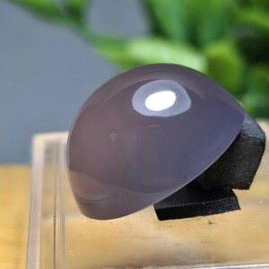 May include: A polished, oval-shaped, lavender-gray gemstone. The stone is mounted on a small, black, rectangular base, and is displayed on a clear, rectangular surface. The background features blurred green foliage.