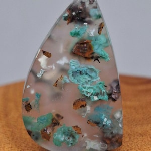 May include: A teardrop-shaped gemstone with a translucent, light pink base. The stone features inclusions of teal, brown, and black mineral deposits. The polished surface is smooth, and the stone is mounted on a black base.