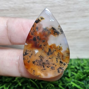 Könnte beinhalten: Ein polierter, tropfenförmiger Edelstein. Der Stein zeigt eine Mischung aus weißen, orangefarbenen und schwarzen Farbtönen mit detaillierten Mustern, die an eine natürliche Landschaft erinnern. Der Edelstein wird zwischen den Fingern gehalten, was seine Form und Größe hervorhebt.