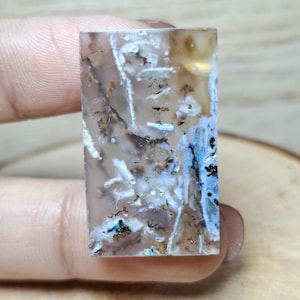 May include: A rectangular gemstone with a translucent appearance. The stone features a mix of colors, including white, brown, and blue, with intricate patterns and inclusions. The gemstone is held between fingers.