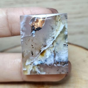 以下が含まれることがあります： 透明なベースに、白、茶色、黒の内部模様が施された長方形の宝石。石は研磨された表面を持ち、自然な外観をしています。宝石は手で持たれています。