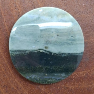 Puede incluir: Una gema redonda y pulida con una apariencia en capas. La parte superior es de un verde claro y nublado, que pasa a un verde bosque más oscuro en la parte inferior. La piedra tiene una superficie lisa y reflectante.