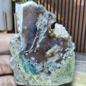 Peut inclure: Une tranche d'agate polie présentant un centre translucide brun et gris avec un bord cristallin irrégulier. La pierre affiche une inclusion mousseuse vert vif à la base, contrastée par une fine veine métallique de couleur cuivre.