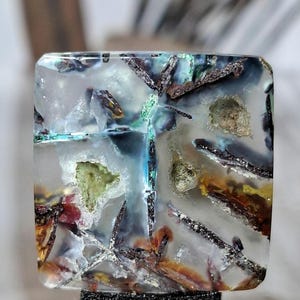 以下が含まれることがあります： 透明な樹脂製の正方形のカボション。内部には鉱物のインクルージョン、暗い金属のような破片、緑、黄、赤褐色の有機的な質感の斑点が埋め込まれており、全体がクリアで光沢のある滑らかな素材で覆われています。
