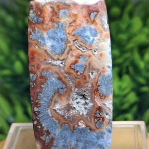 May include: A rectangular gemstone with a polished surface. The stone features a complex pattern of blue, orange, and white swirls and inclusions. The gemstone is set on a clear display stand, showcasing its natural beauty.