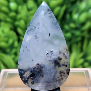 Puede incluir: Una gema en forma de lágrima con una superficie pulida. La piedra presenta una base blanca translúcida con intrincados patrones de inclusiones negras y grises, creando una apariencia nublada y natural. La gema está colocada sobre una base negra.