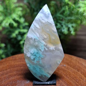 以下が含まれることがあります： ユニークな模様の、磨かれた涙型の宝石。 白、黄色、ターコイズの渦巻き模様があり、雲や風景を連想させます。 小さな黒い台座に取り付けられており、その自然な美しさを際立たせています。