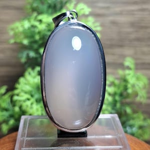May include: Oval-shaped pendant with a pale gray gemstone set in a silver-colored metal frame. The pendant has a smooth, polished surface and a small loop at the top for a chain. The background features green foliage.