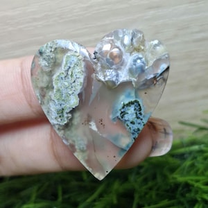 May include: A heart-shaped gemstone with a translucent base, featuring green, blue, and white inclusions. The stone has a polished surface and a natural, organic appearance. The gemstone is a unique piece of jewelry.