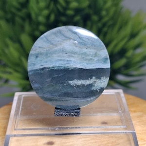 Puede incluir: Una gema redonda y pulida con un patrón en capas de azul, verde y blanco. La piedra está montada sobre un soporte de acrílico transparente. Los colores y patrones naturales recuerdan a un paisaje.
