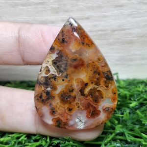 Könnte beinhalten: Ein polierter, tropfenförmiger Edelstein. Der Stein zeigt eine Mischung aus orangefarbenen, braunen und schwarzen Mustern, die an eine Landschaft erinnern. Die Grundfarbe ist cremeweiß, mit dunkleren Flecken und Adern. Der Edelstein wird von einer Hand gehalten.